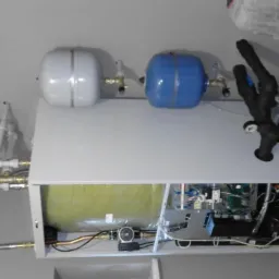 Instalacja z zbiornikami wyrównawczymi: białym i niebieskim, zaworami, rurami miedzianymi i czarnymi, manometrem, widoczne wewnętrzne okablowanie i elektronika urządzenia.
