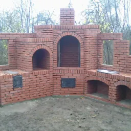 Murowana z czerwonej cegły konstrukcja grilla ogrodowego z trzema piecami, kominem i blatem roboczym, na tle drzew bez liści.