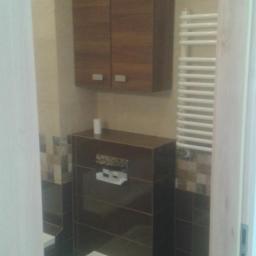 Michal sasiela - Nowoczesna łazienka z podwieszaną toaletą, zabudową z ciemnych płytek i szafką wiszącą z drewnianymi frontami, uzupełniona o biały grzejnik drabinkowy.