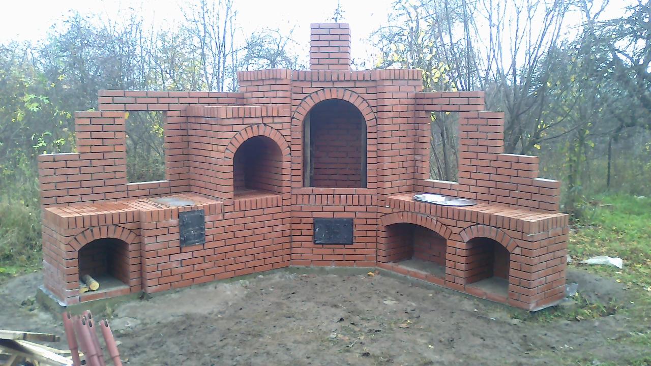 Murowana z czerwonej cegły konstrukcja grilla ogrodowego z trzema piecami, kominem i blatem roboczym, na tle drzew bez liści.