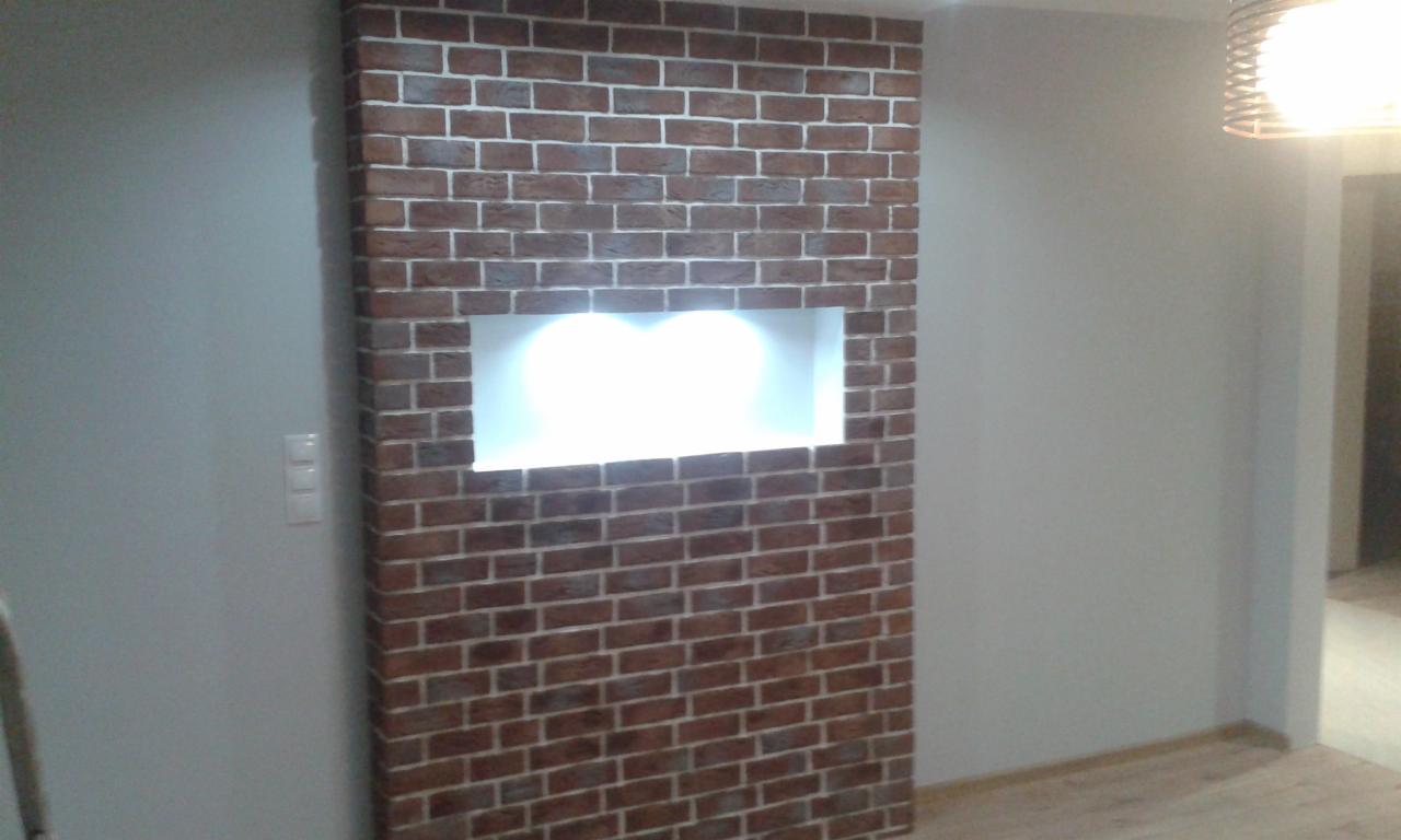 Ściana w salonie wykończona cegłą klinkierową z podświetloną wnęką, widoczny fragment parkietu i lampy wiszącej.
