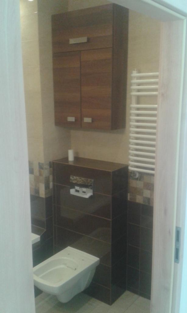 Nowoczesna łazienka z podwieszaną toaletą, zabudową z ciemnych płytek i szafką wiszącą z drewnianymi frontami, uzupełniona o biały grzejnik drabinkowy.