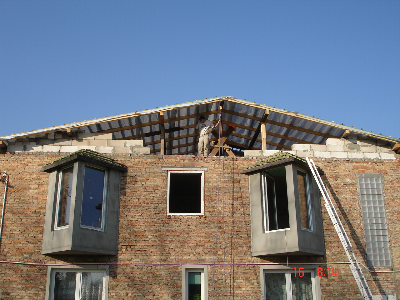 Częściowo zdemontowany dach budynku z czerwonej cegły, widoczna konstrukcja drewniana i folia, dekarz na dachu zabezpieczony liną, drabina oparta o ścianę.