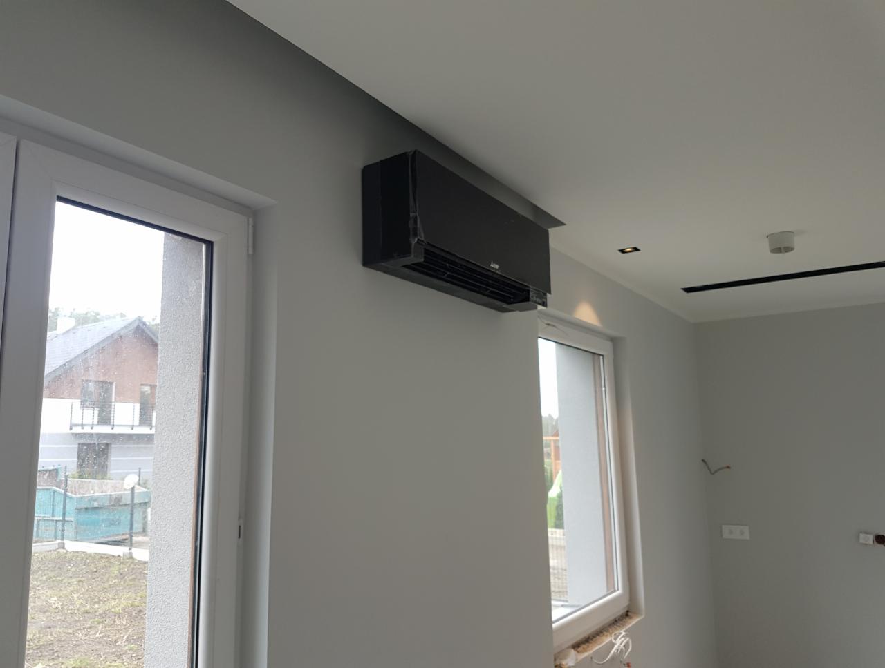 Czarny klimatyzator ścienny Mitsubishi Electric zamontowany w szarym pokoju z dwoma oknami, widok z dołu.