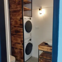 Nowoczesna łazienka z pralko-suszarką w zabudowie z ciemnego drewna i metalu, umywalka nablatowa na drewnianej szafce, czarno-biała podłoga w geometryczny wzór.