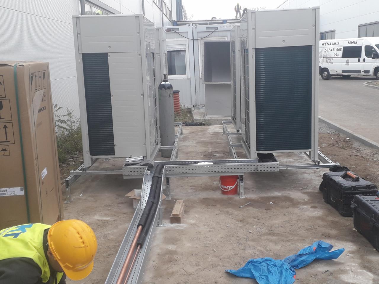 Instalacja zewnętrznych jednostek klimatyzacyjnych na metalowych konstrukcjach, widoczne rury miedziane w korytkach kablowych, butla z gazem technicznym oraz pracownik w żółtym kasku