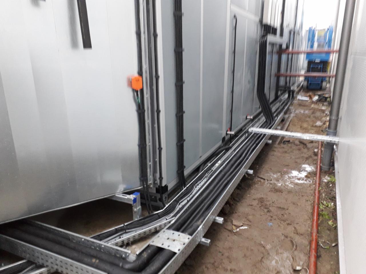 Instalacja przewodów wentylacyjnych i elektrycznych na metalowych korytach wzdłuż ściany budynku przemysłowego, widoczny podnośnik w tle.