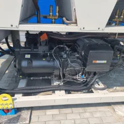 Agregat chłodniczy na zewnątrz budynku, widoczne rury, zawory i sprężarka. Elementy konstrukcyjne pomalowane na biało, całość ustawiona na kostce brukowej.