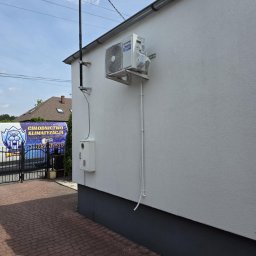 Cold ice klim - Biały klimatyzator zamontowany na elewacji budynku, z widocznymi przewodami i skrzynką elektryczną. W tle brama i samochód firmy chłodniczej.