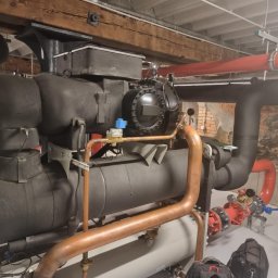 Cold ice klim - Duża przemysłowa instalacja chłodnicza z rurami miedzianymi i czerwonymi, zamontowana w pomieszczeniu z ceglanym murem w tle.