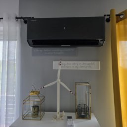 Cold ice klim - Czarny klimatyzator Hisense na ścianie w pokoju z dekoracjami, widoczny pilot. Żółte i białe zasłony po bokach. Widok z okna na budynki w tle.