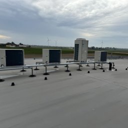 Cold ice klim - Instalacja klimatyzacji na dachu budynku. Widoczne jednostki zewnętrzne na metalowej konstrukcji wsporczej. W tle krajobraz z turbinami wiatrowymi.