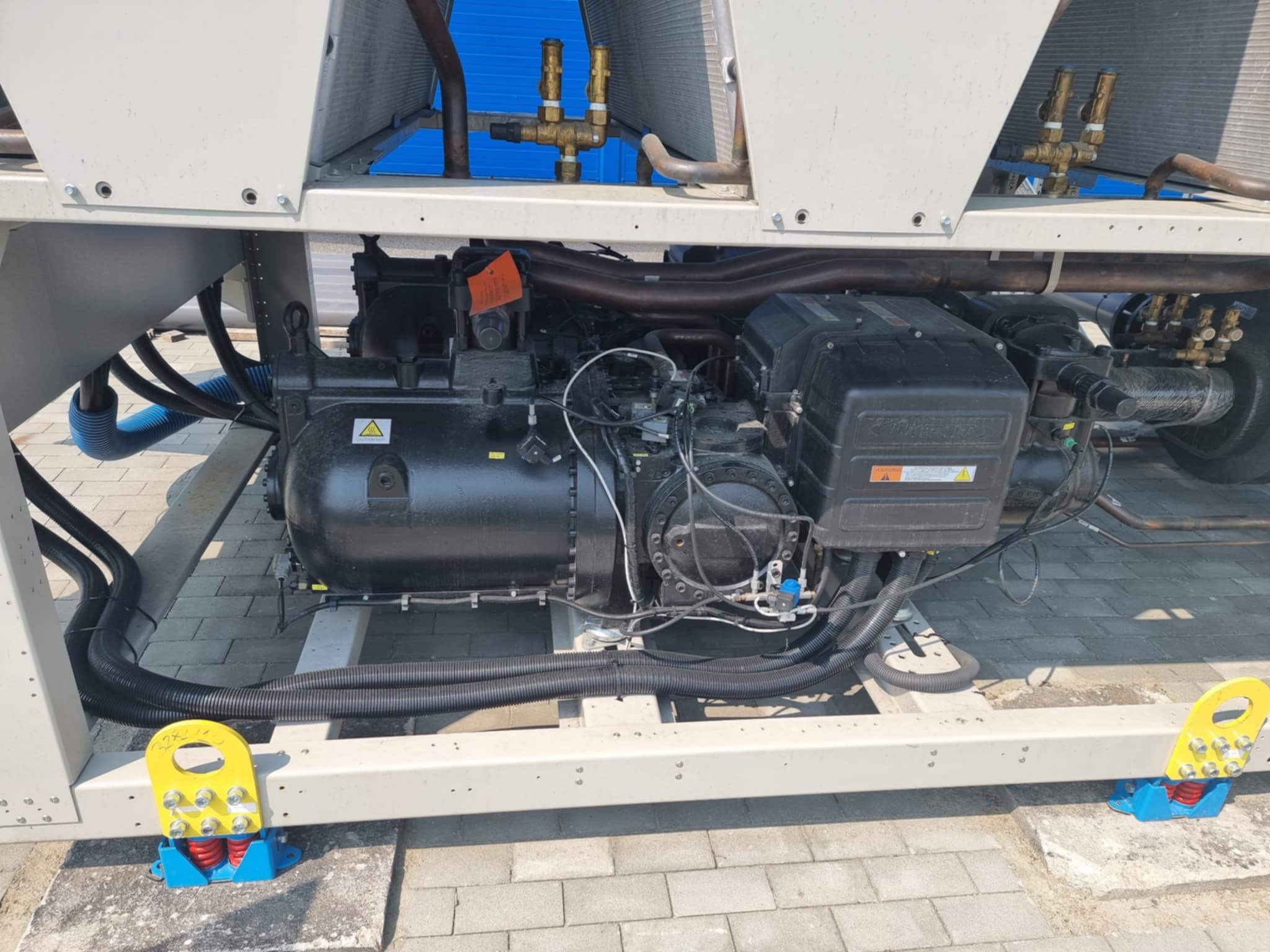 Agregat chłodniczy na zewnątrz budynku, widoczne rury, zawory i sprężarka. Elementy konstrukcyjne pomalowane na biało, całość ustawiona na kostce brukowej.
