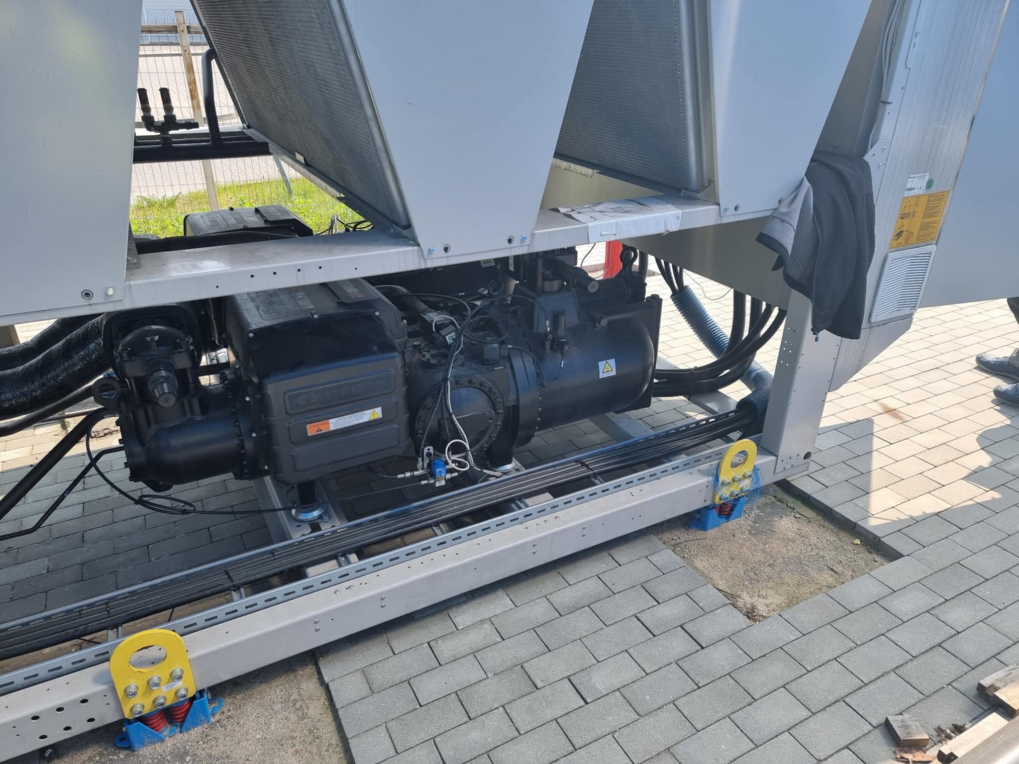 Agregat chłodniczy na zewnątrz budynku, zamontowany na metalowej konstrukcji z amortyzatorami. Widoczne przewody i elementy instalacji. Tło stanowią kostki brukowe.