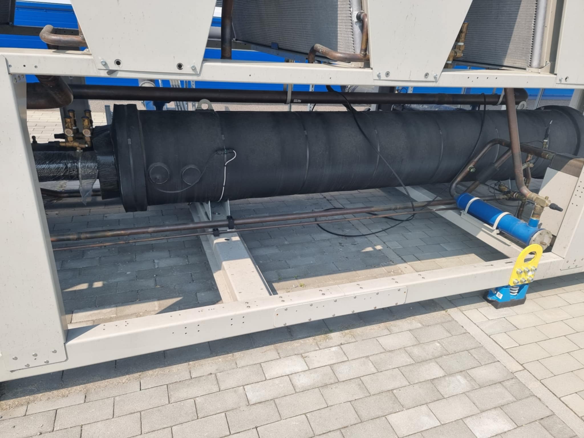 Agregat chłodniczy z rurami w izolacji, widoczne elementy mocujące i konstrukcja wsporcza. Urządzenie stoi na kostce brukowej.