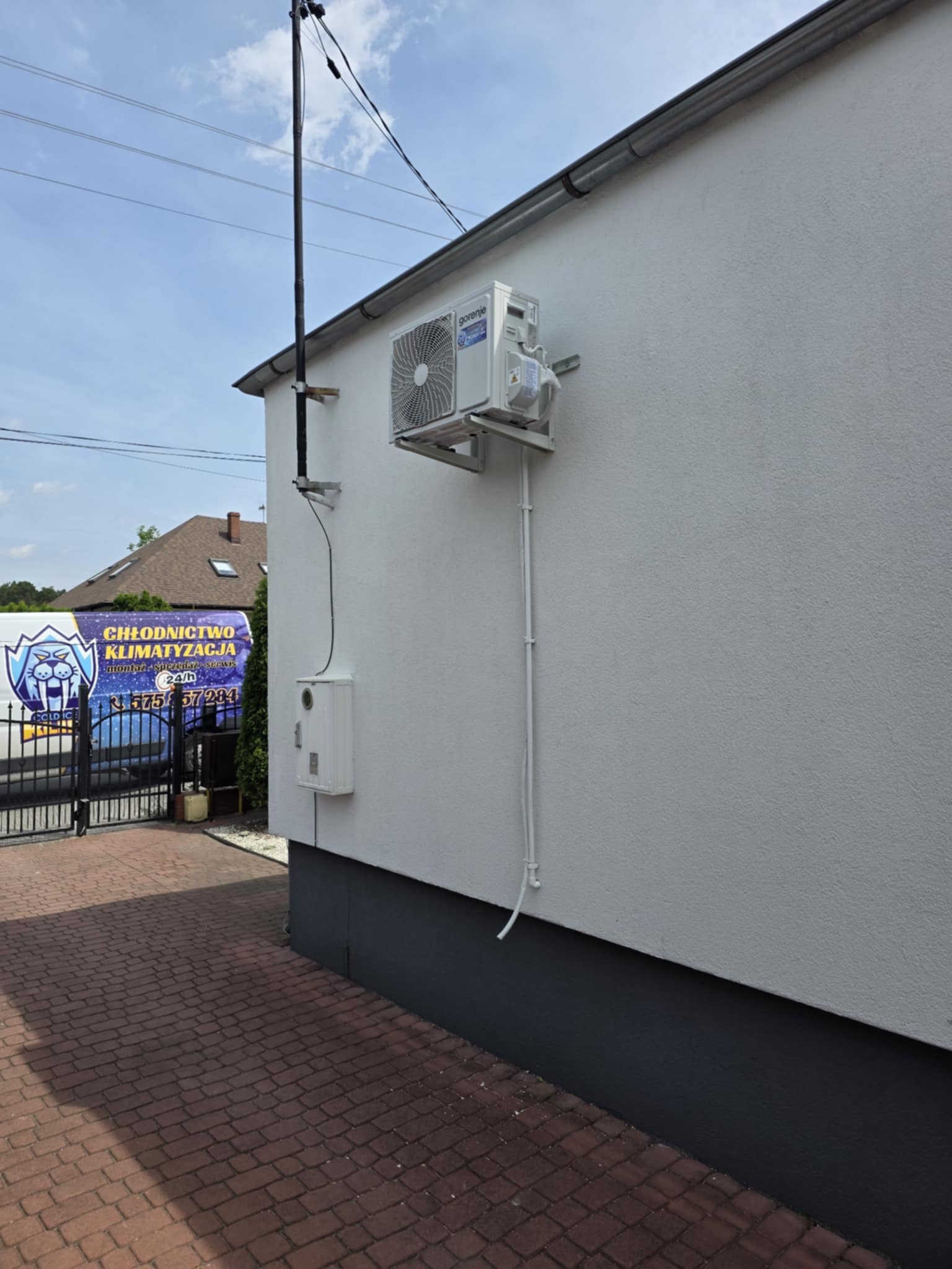 Biały klimatyzator zamontowany na elewacji budynku, z widocznymi przewodami i skrzynką elektryczną. W tle brama i samochód firmy chłodniczej.