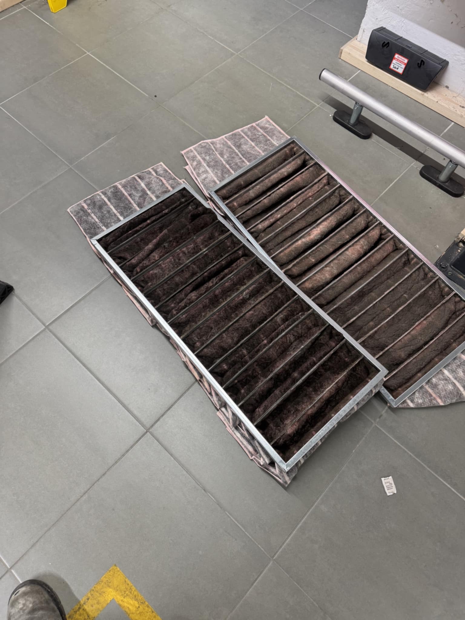 Dwa zużyte, prostokątne filtry klimatyzacyjne z metalową ramą, leżące na szarej podłodze z płytek. Widoczne zabrudzenia i włókna wewnątrz filtrów. Filtracja powietrza.