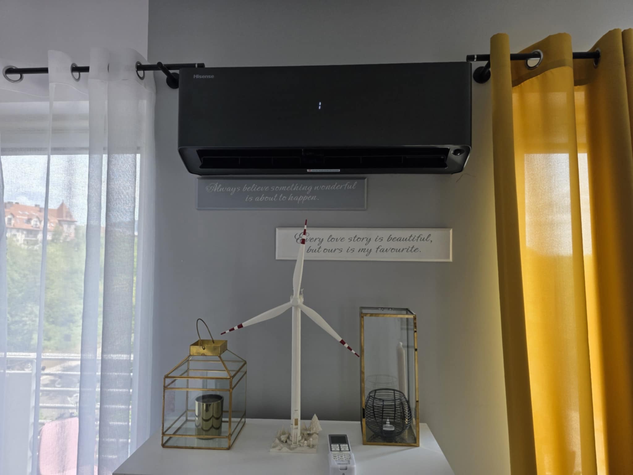 Czarny klimatyzator Hisense na ścianie w pokoju z dekoracjami, widoczny pilot. Żółte i białe zasłony po bokach. Widok z okna na budynki w tle.