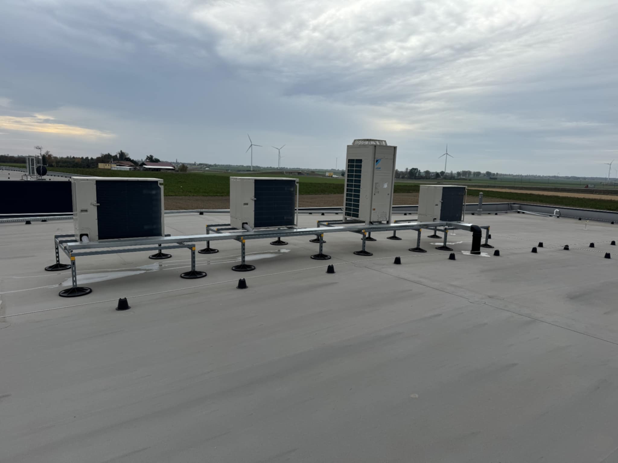 Instalacja klimatyzacji na dachu budynku. Widoczne jednostki zewnętrzne na metalowej konstrukcji wsporczej. W tle krajobraz z turbinami wiatrowymi.