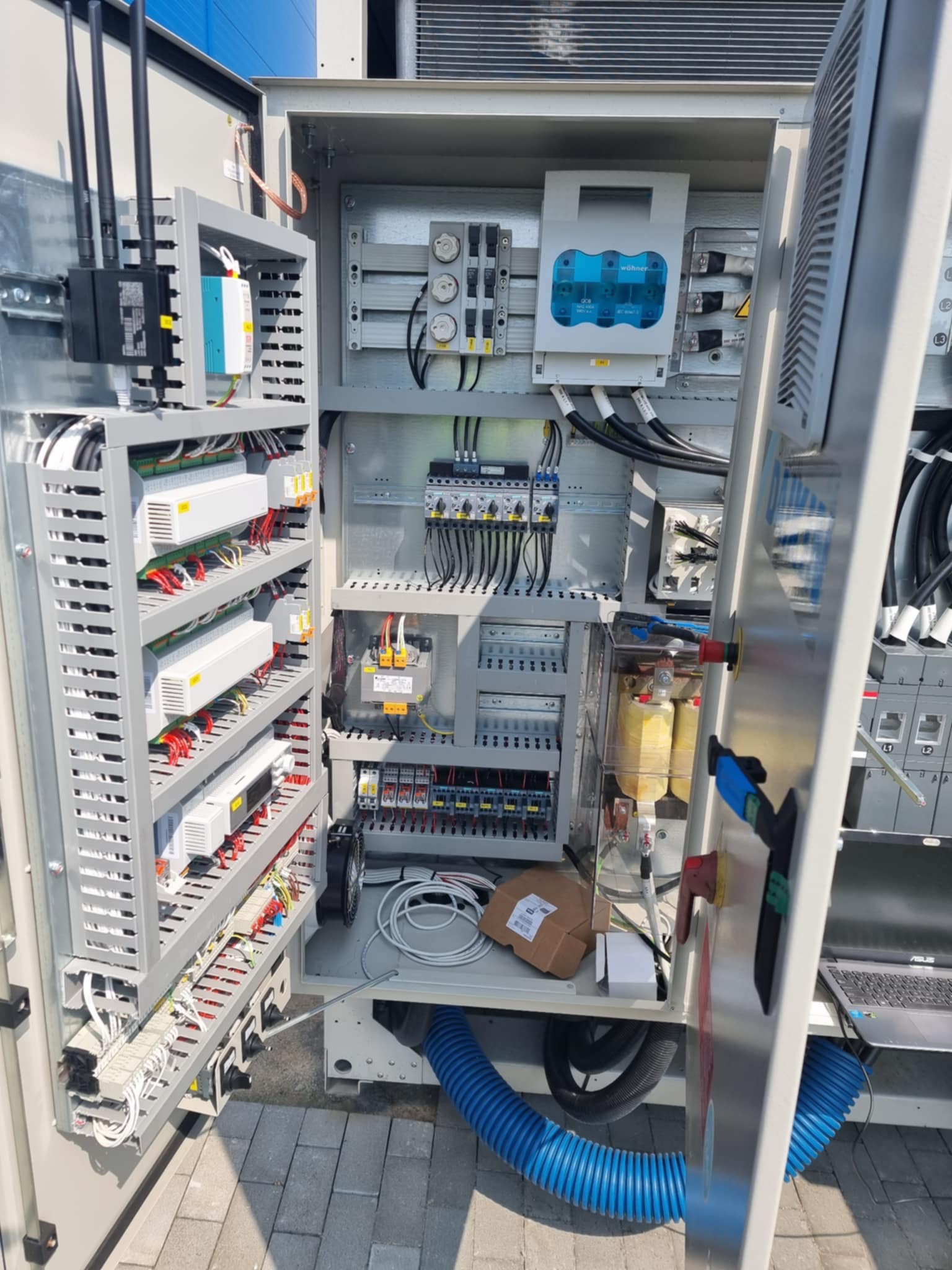 Otwarta szafa sterownicza z modułami, okablowaniem i elementami automatyki. Widoczny laptop na półce, sugerujący konfigurację lub diagnostykę systemu.