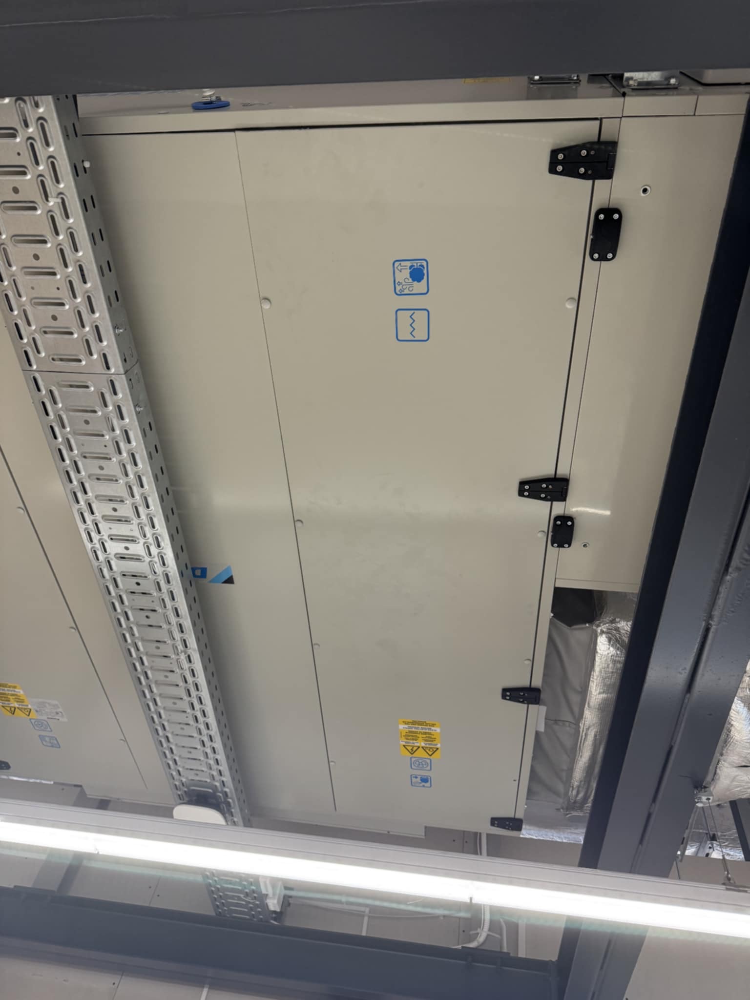 Sufitowa instalacja klimatyzacyjna z widocznymi panelami dostępowymi, kanałami wentylacyjnymi i elementami konstrukcyjnymi. Widok z dołu, oświetlenie jarzeniowe.