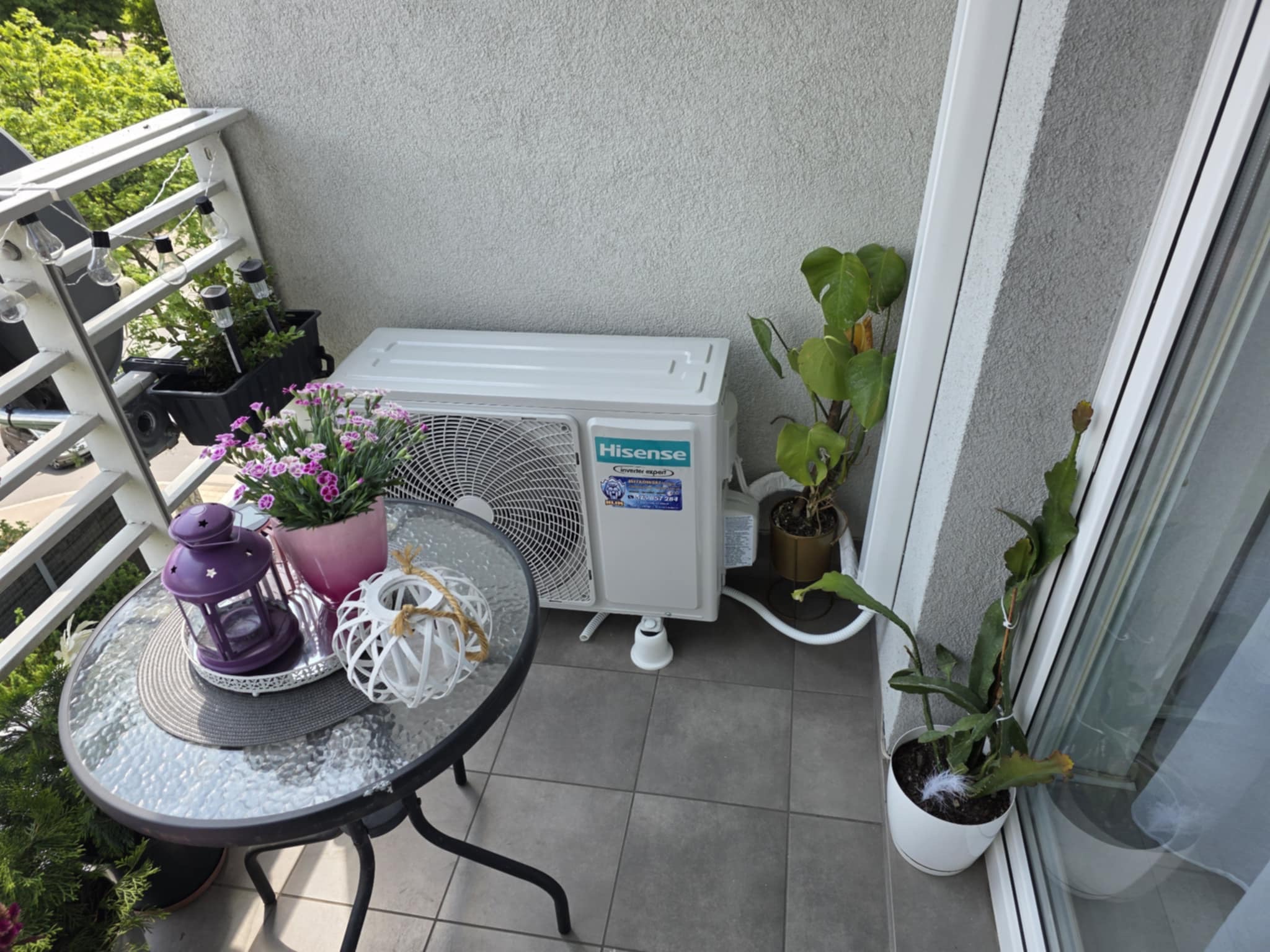 Biały klimatyzator Hisense na balkonie z kwiatami, lampionem i roślinami doniczkowymi, tworzący przytulny kącik.