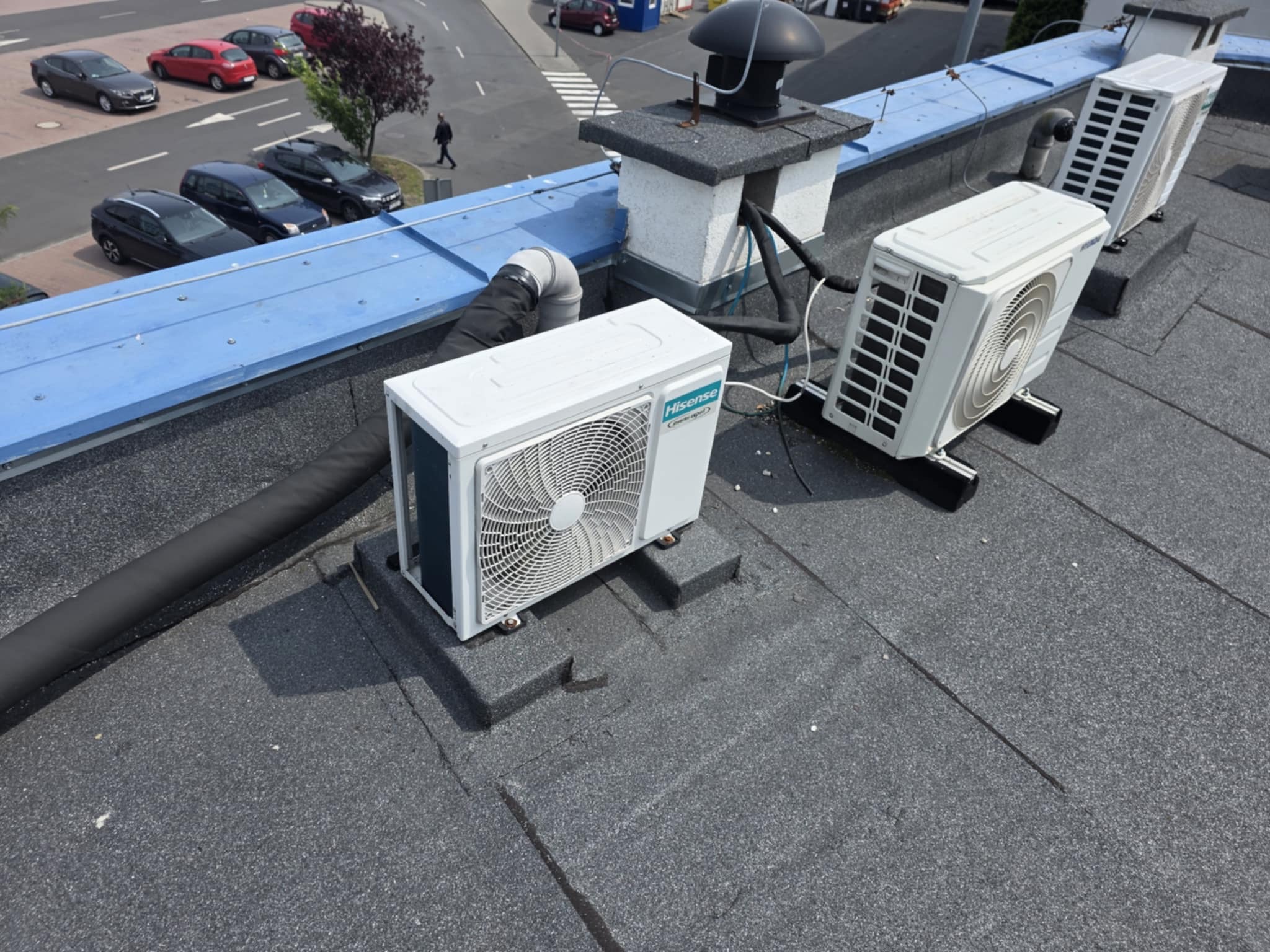 Białe jednostki klimatyzacji Hisense na płaskim dachu budynku, widok z góry na ulicę z samochodami i pieszym. Instalacja klimatyzacji na dachu.