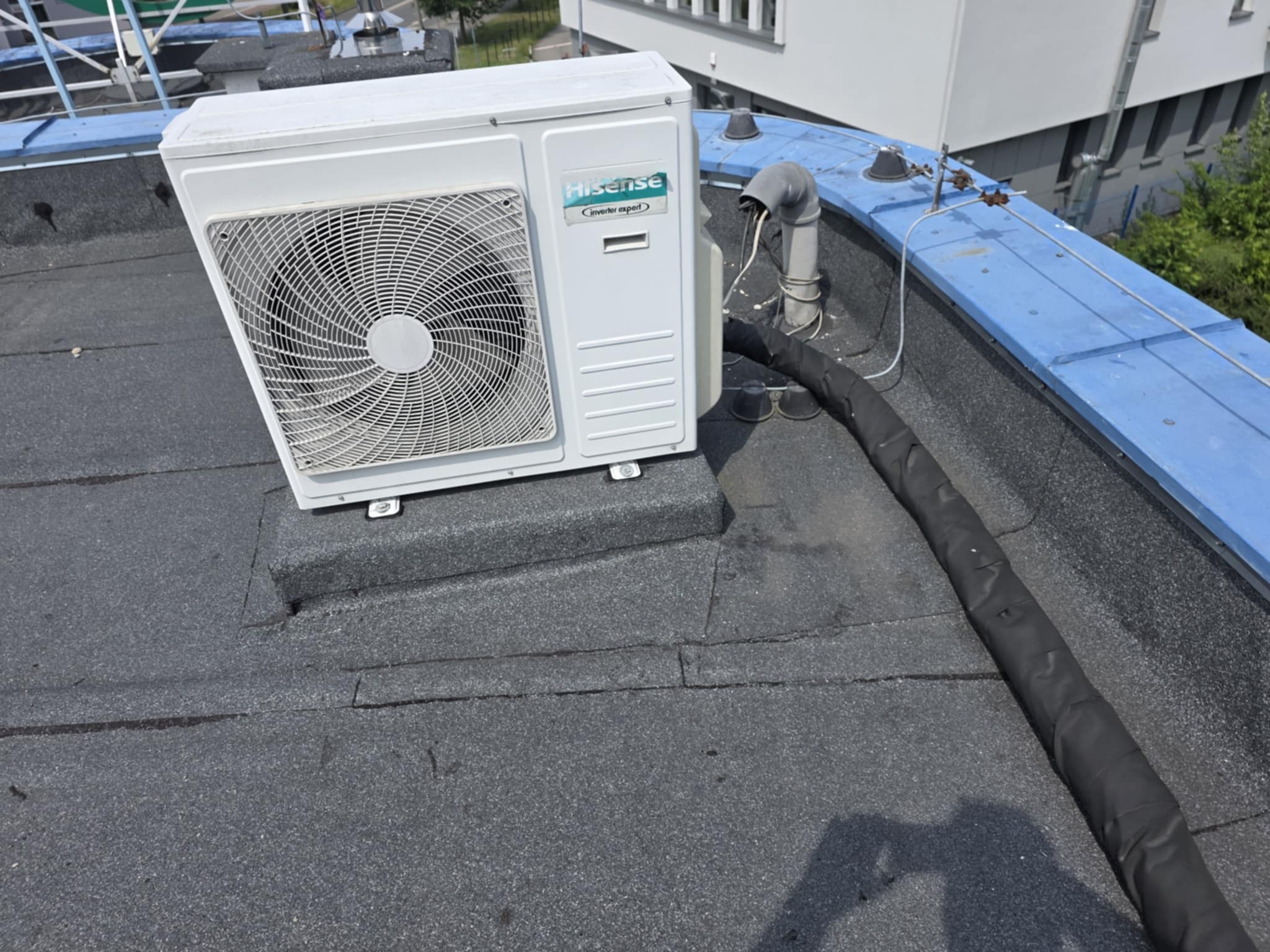 Biała jednostka zewnętrzna klimatyzacji Hisense na czarnym, płaskim dachu z widocznymi przewodami i elementami konstrukcji budynku w tle.