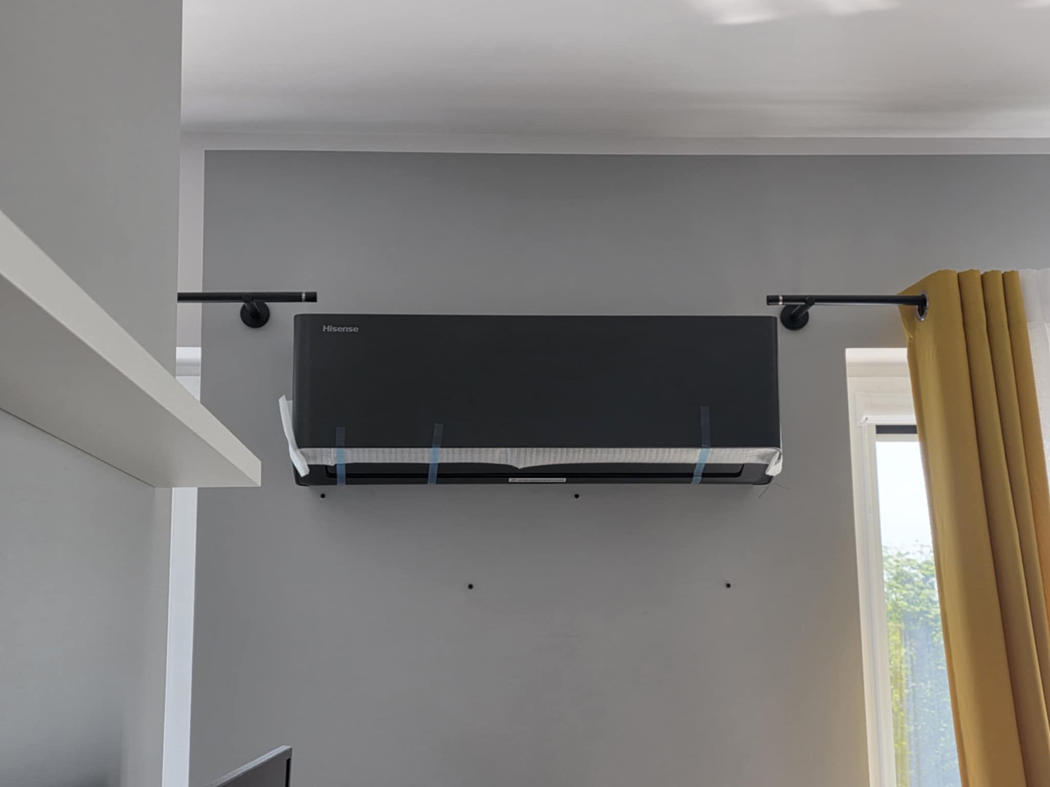 Czarny klimatyzator Hisense zamontowany na szarej ścianie, zabezpieczony folią i taśmą montażową. Widoczne elementy wystroju wnętrza: karnisz i zasłona.