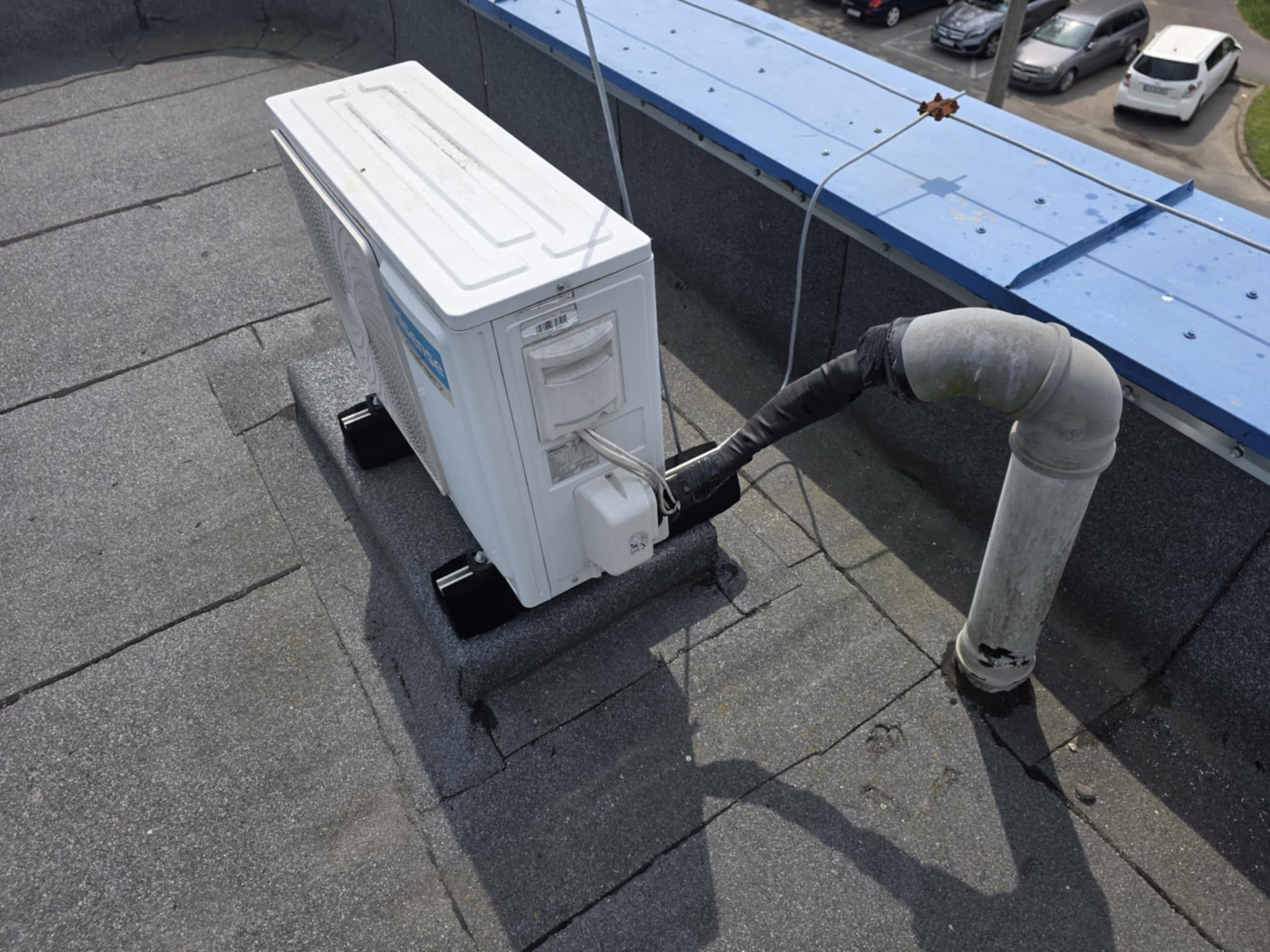 Biała jednostka zewnętrzna klimatyzacji na płaskim dachu, z widocznymi przewodami i rurą odprowadzającą skropliny. W tle zaparkowane samochody.