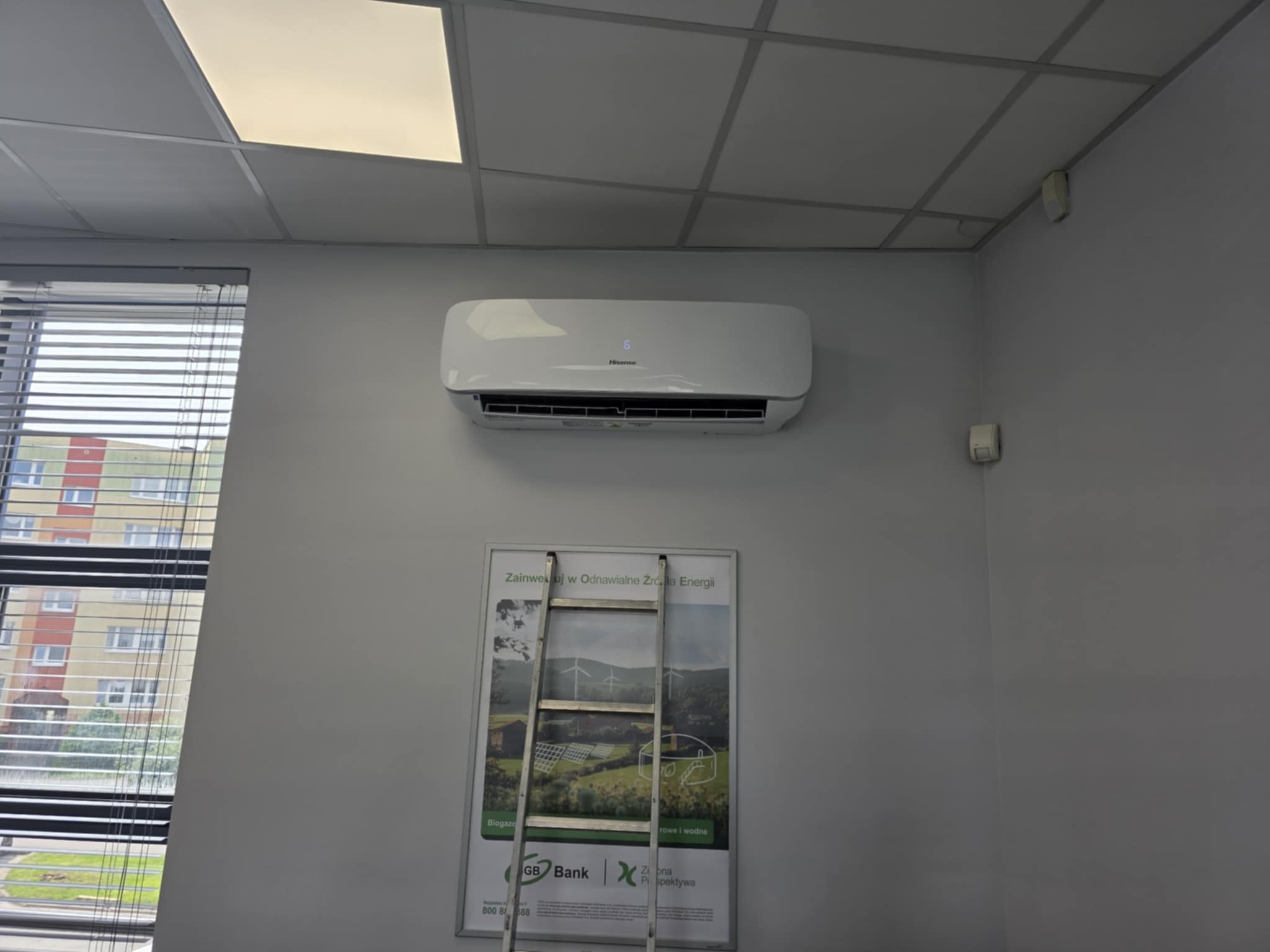 Biała klimatyzacja Hisense na szarej ścianie biura, nad plakatem 'Zainwestuj w Odnawialne Źródła Energii' i drabiną opartą o niego. Widok z dołu.