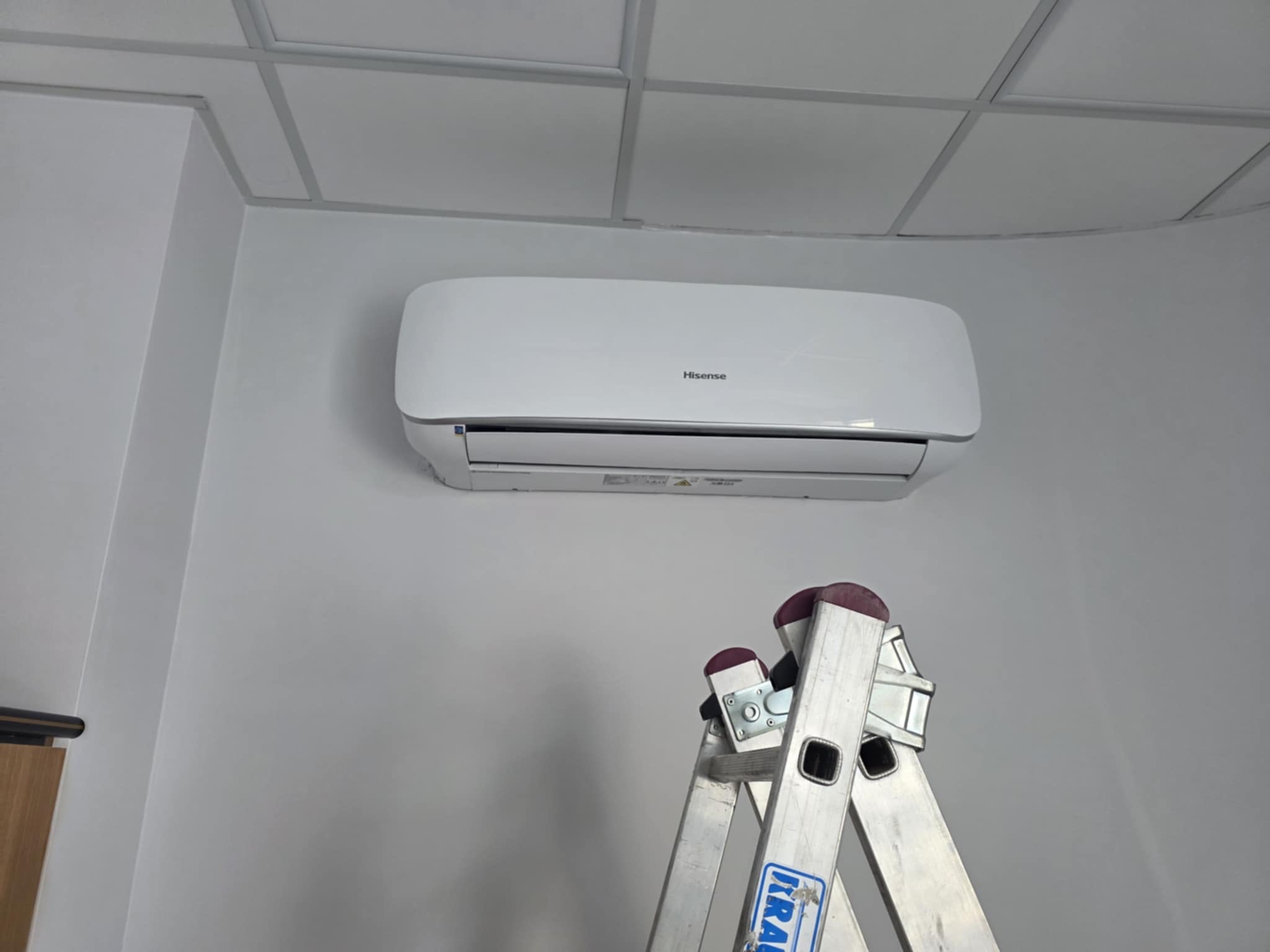 Biały klimatyzator Hisense na ścianie biura, widoczna drabina aluminiowa. Montaż klimatyzacji w pomieszczeniu z sufitem kasetonowym.