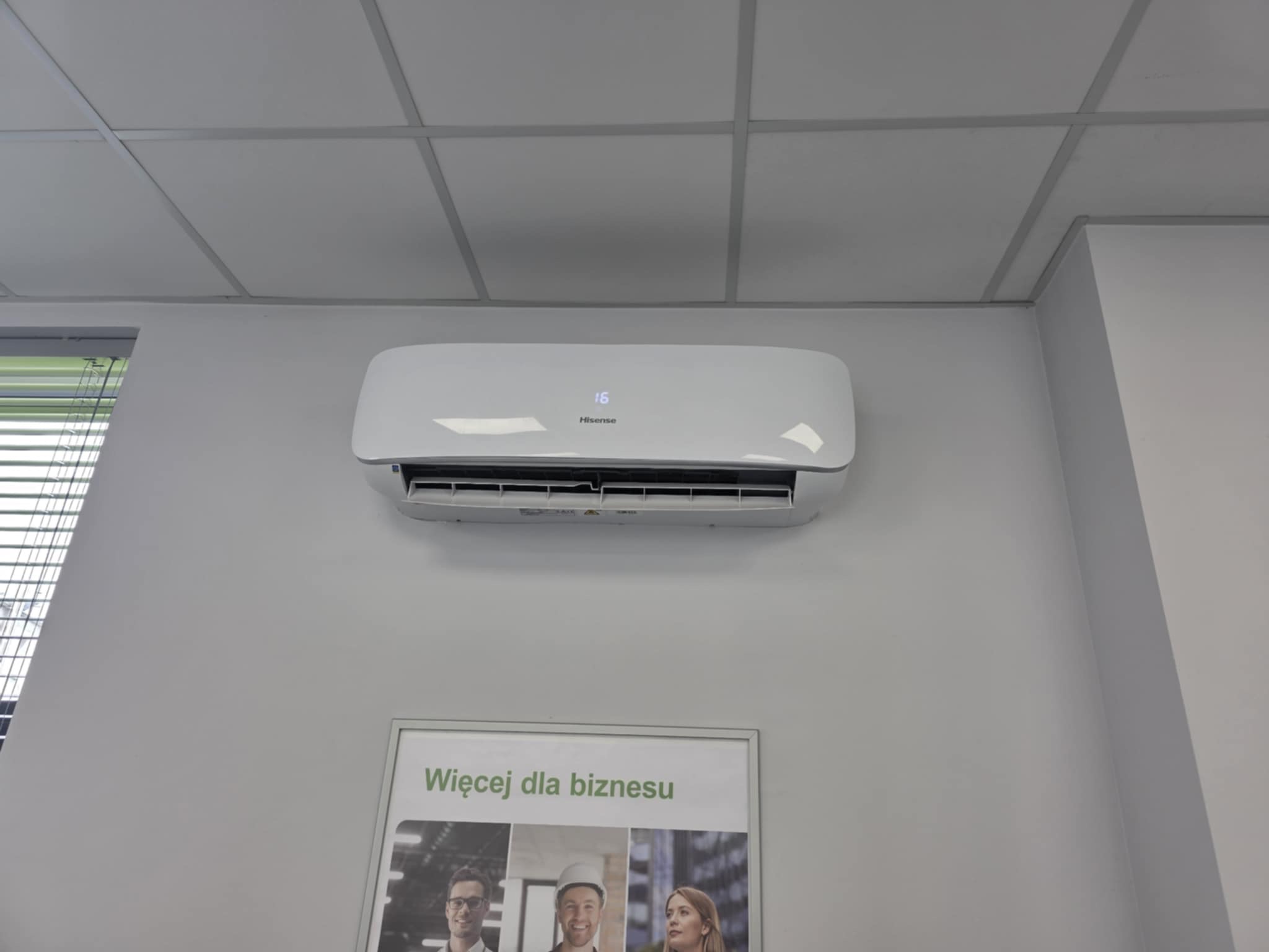 Biała klimatyzacja Hisense zamontowana na ścianie biura, pod sufitem kasetonowym. Pod klimatyzatorem widoczny plakat reklamowy.