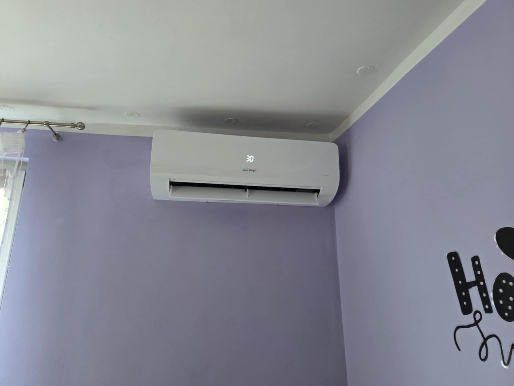 Biały klimatyzator Gorenje z wyświetlaczem 30 stopni Celsjusza zamontowany na liliowej ścianie w pomieszczeniu. Widoczny fragment dekoracji ściennej.