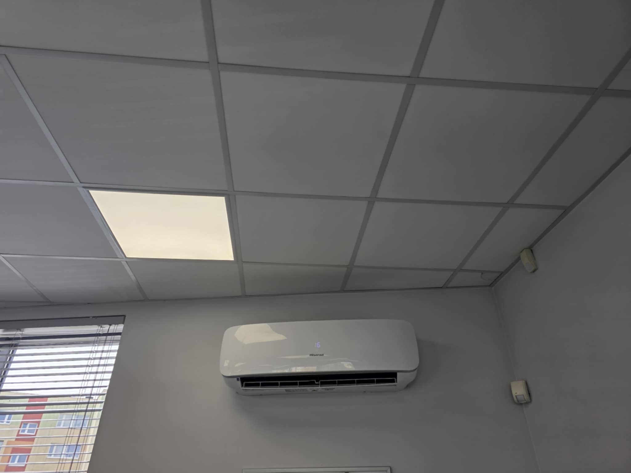 Biała klimatyzacja Hisense na szarej ścianie biura, widok z dołu, z żaluzjami i kasetonowym sufitem. Delikatne oświetlenie.
