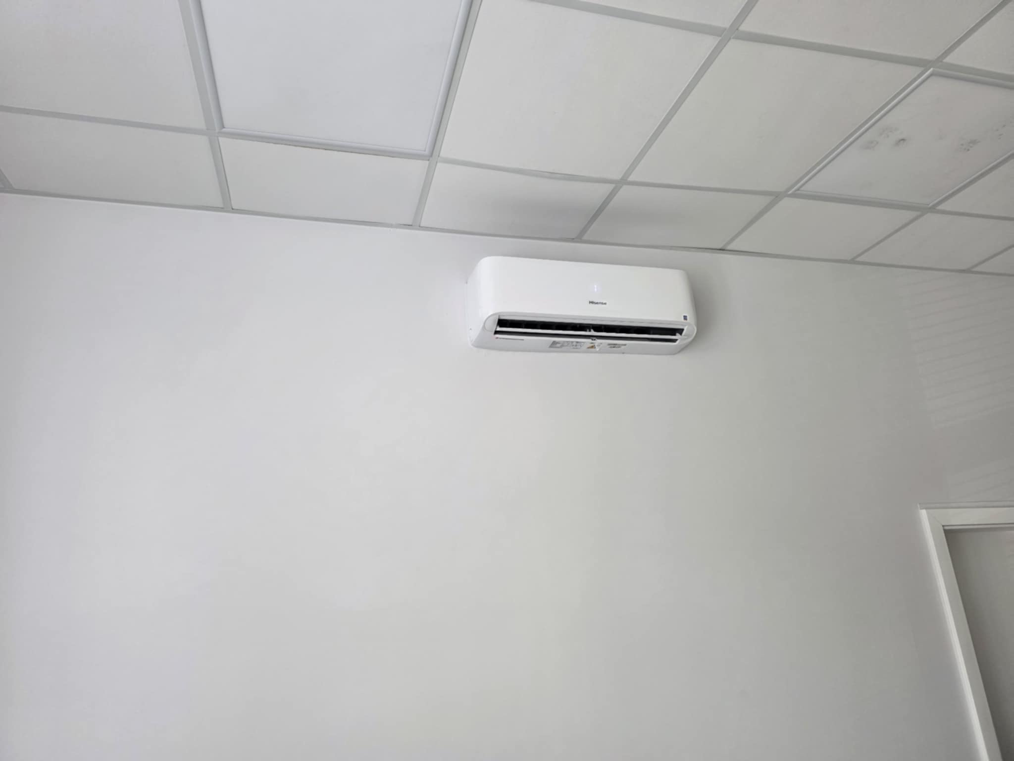 Biała klimatyzacja Hisense zamontowana na jasnej ścianie pod sufitem kasetonowym, widoczna z boku, w tle fragment futryny drzwi.