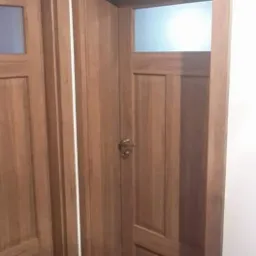 Dwa skrzydła z drewna z szybami w górnej części, zamontowane w jasnym pomieszczeniu, widoczne klamki i faktura drewna.