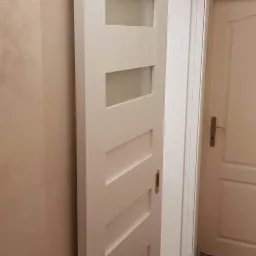 Białe, częściowo przeszklone skrzydło przesuwne z poziomymi panelami, zamontowane w systemie szynowym, widoczne na tle beżowej ściany i brązowych płytek podłogowych.