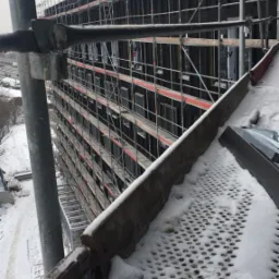 Wielopiętrowy budynek w trakcie ocieplania, osłonięty rusztowaniami w zimowej scenerii z widocznym śniegiem na rusztowaniach.