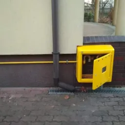 Żółta skrzynka gazowa z otwartymi drzwiczkami, zamontowana na ceglanym cokole budynku, z widocznymi rurami gazowymi wewnątrz.