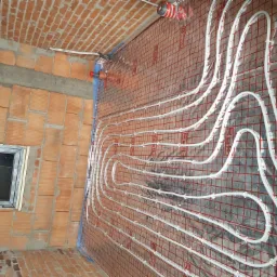 Instalacja ogrzewania podłogowego w surowym wnętrzu z ceglanymi ścianami i oknem, rury układane na siatce montażowej, widoczne elementy przyłączeniowe.