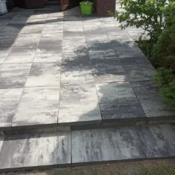 Ułożona kostka brukowa w odcieniach szarości prowadząca do wejścia do domu, z widocznymi dwoma stopniami i zielenią po prawej stronie.