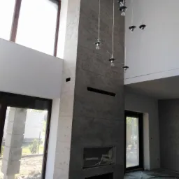 Wysoki komin z tynkowaną elewacją wewnętrzną w surowym stylu, widoczne okna i prowizoryczne oświetlenie, podłoga zabezpieczona folią malarską.