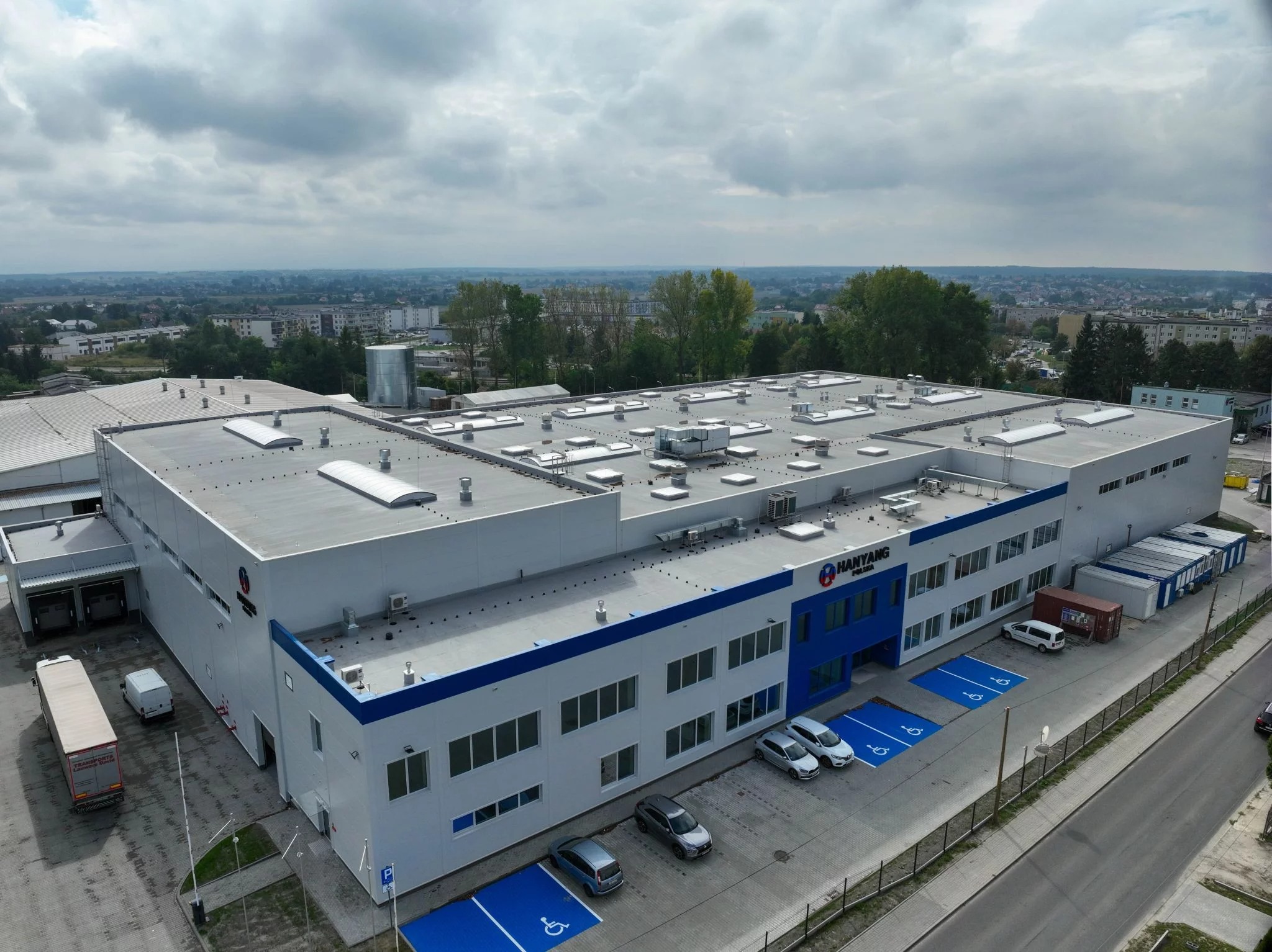 Szeroki kadr z lotu ptaka na nowoczesny budynek przemysłowy z szarym dachem, niebieskimi akcentami i parkingiem. Widoczne samochody i ciężarówka. Pochmurne niebo w tle.