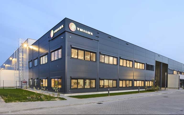 Nowoczesny budynek biurowy z szarej blachy z oknami i logo Trulux na dachu, oświetlony od wewnątrz, z chodnikiem i zielenią na pierwszym planie.