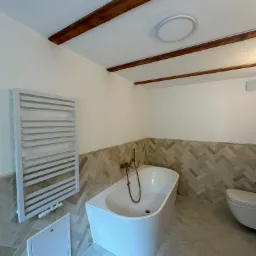 Biała, wolnostojąca wanna z mosiężną armaturą w nowoczesnej łazience z drewnianymi belkami na suficie i grzejnikiem drabinkowym. Ściany wyłożone płytkami w jodełkę.