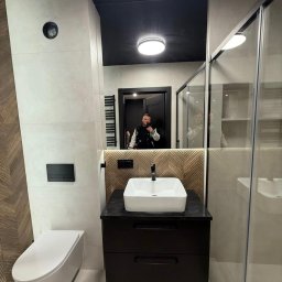 KARLITO WYKOŃCZENIA WNĘTRZ ADRIAN GŁUSZCZAK - Nowoczesna łazienka z białą ceramiką, czarnymi akcentami i drewnianą mozaiką na ścianie, odbicie osoby robiącej zdjęcie w lustrze.