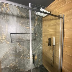 Nowoczesna kabina prysznicowa z przesuwnymi drzwiami, wykończona płytkami imitującymi marmur z złotymi żyłami i drewnopodobnymi, z czarną armaturą i sufitem.