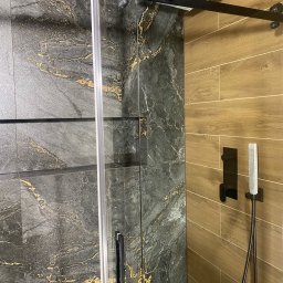 Kabina prysznicowa z przesuwnymi drzwiami, wykończona płytkami imitującymi marmur z złotymi żyłami i drewnopodobnymi panelami. Nowoczesny design, czarne akcenty.