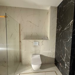 KARLITO WYKOŃCZENIA WNĘTRZ ADRIAN GŁUSZCZAK - Elegancka łazienka po remoncie: białe i czarne marmurowe płytki, kabina prysznicowa ze złotymi detalami, nowoczesna toaleta. Widok na wykończone wnętrze.